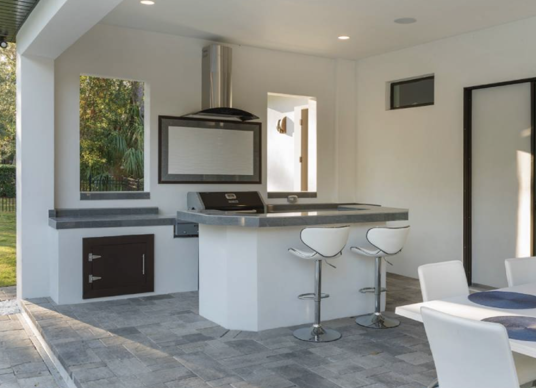 Modern outdoor kitchen with a built-in grill, range hood, two bar stools at a counter, and white walls with large windows.